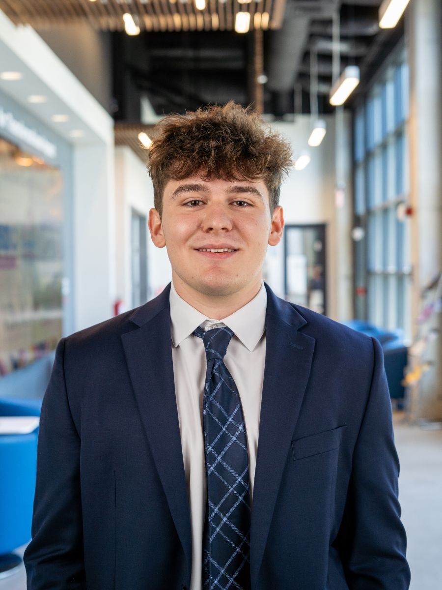 Dennis Tudor poses for a headshot in a blue suit and tie in a bright room filled with windows and lights on the ceiling. 