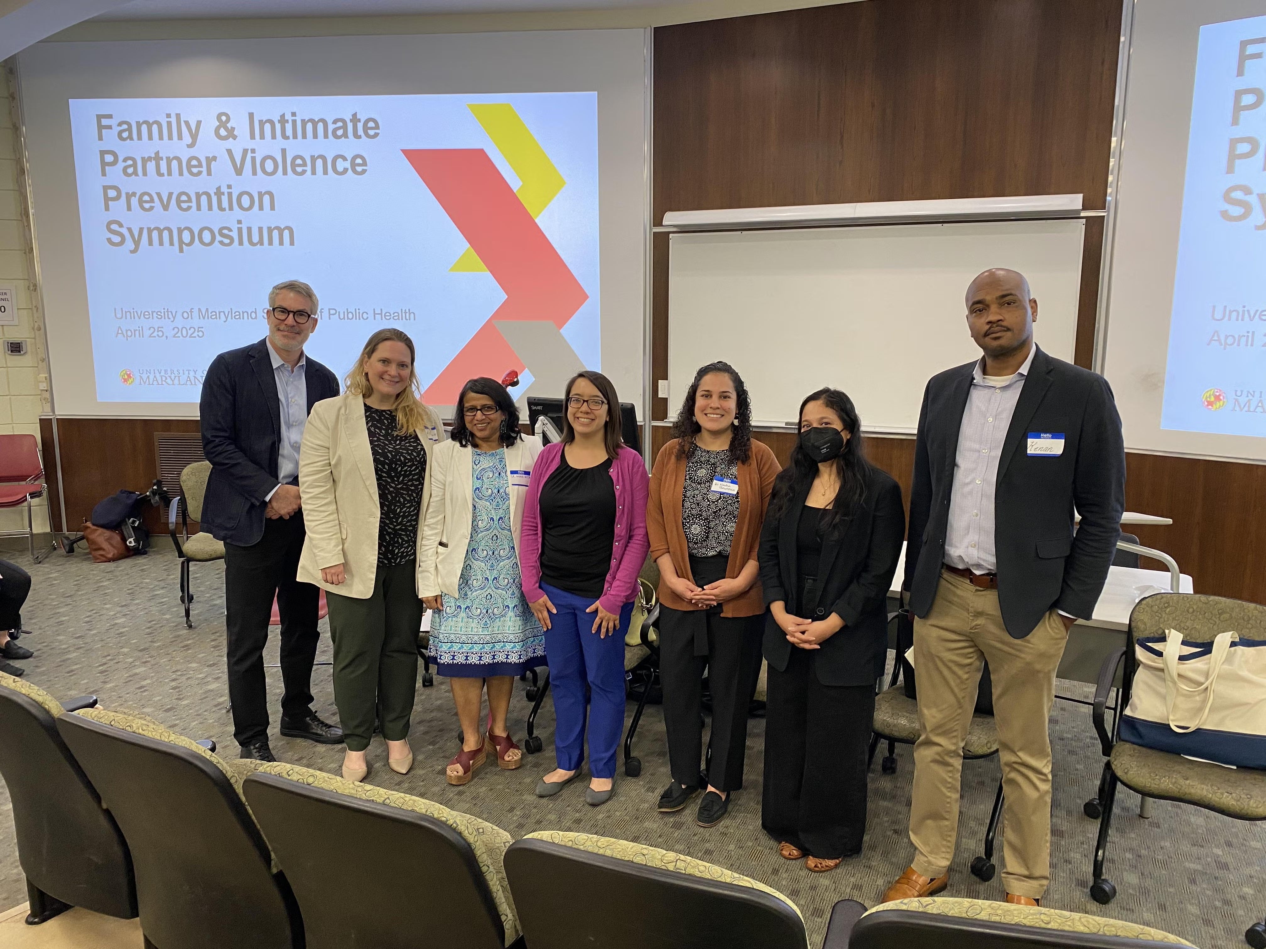A group of speakers posing for a group photo up on the front of a stage in a classroom. 