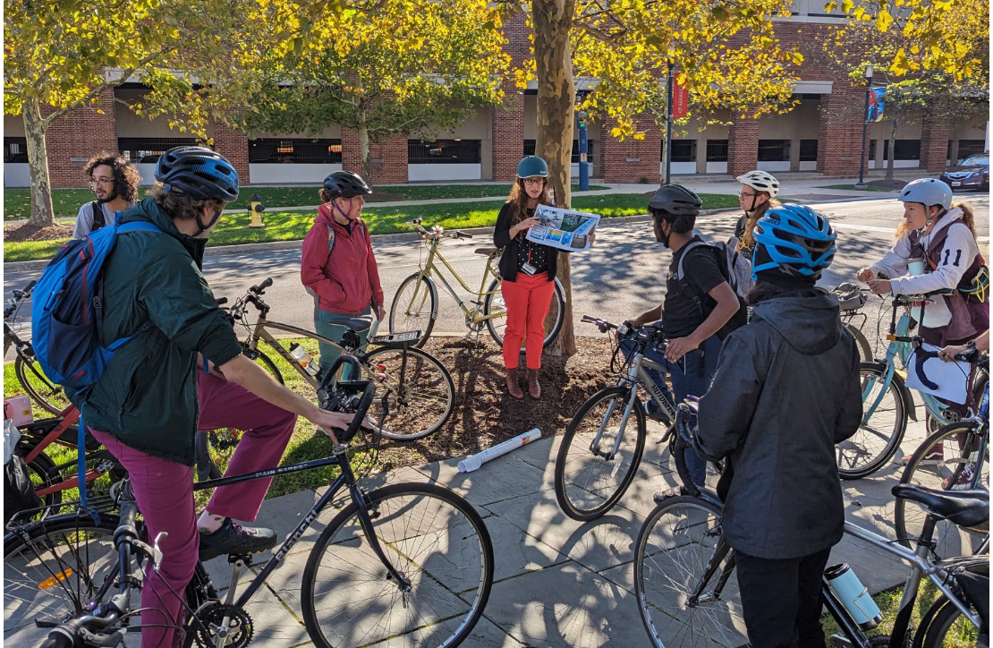 A group on campus wearing bike helmets, standing next to bikes, watching someone hold up a pamphlet and provide information
