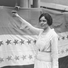 Black and white image of a woman toasting with a glass of wine to a flag with stars on it