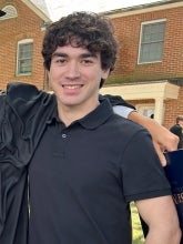 Liam Trump's headshot features him with black hair and a black shirt posed for a phot with a brick building with two windows behind him.