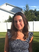 Megan Dougherty's headshot. She stands in front of a white fence with a sunny background filled with green grass and blue skies. 