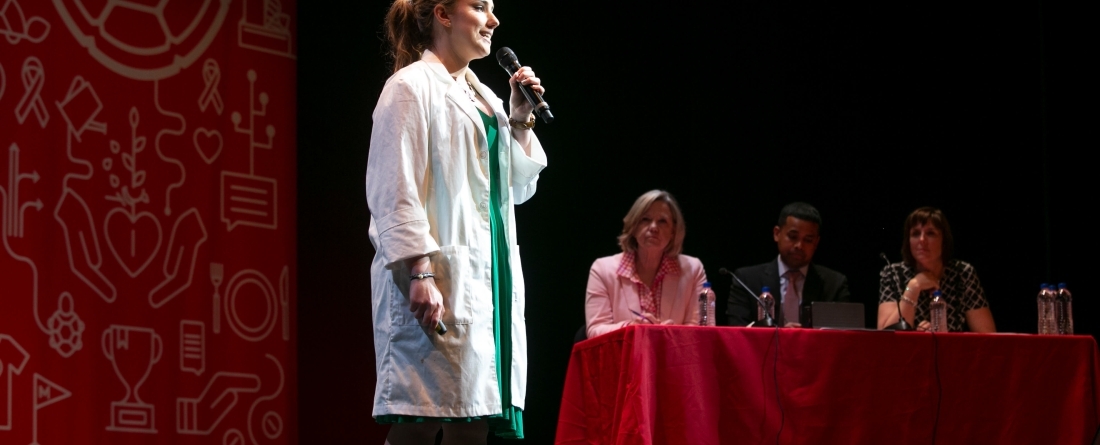 Woman standing on a stage in white lab coat holding a microphone