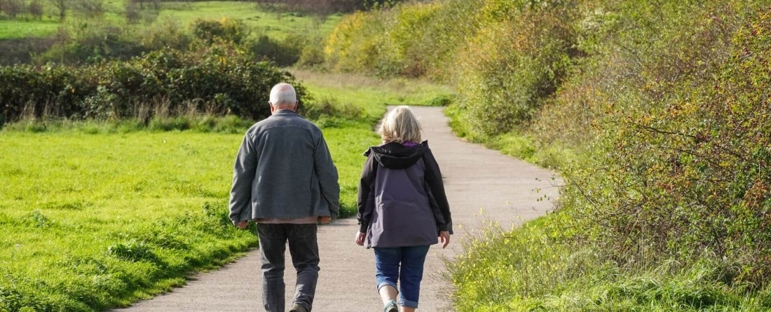 image of 2 older adults from behind walking on a path holding hands surrounded by green grass