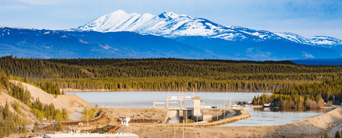 A head-on view of a hydropower dam surrounded by a green forests with a large snow-capped mountain in the background.