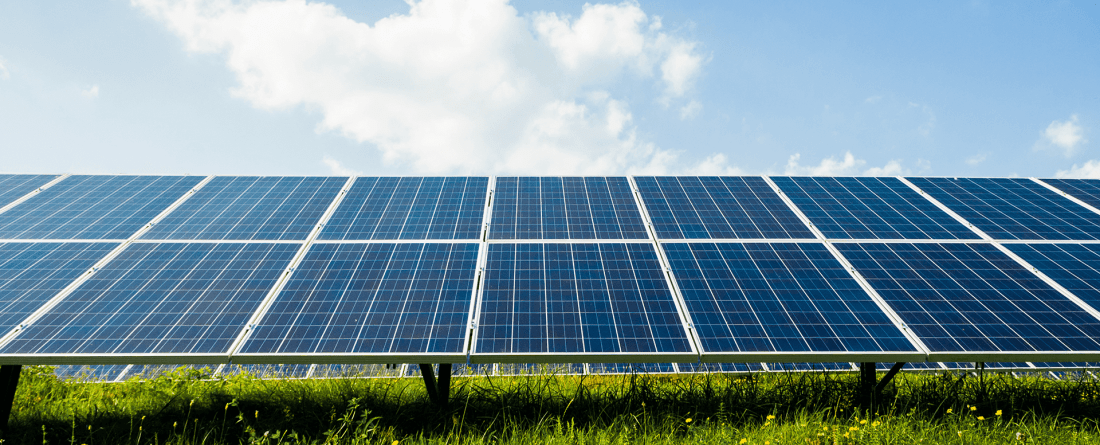 An array of solar panels in a grassy field on a day with blue skies and a few clouds.