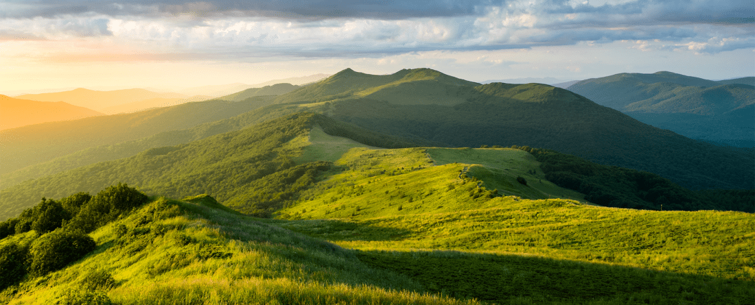 A grassy mountainous area with blue skies and clouds that rest above mountains in the background.