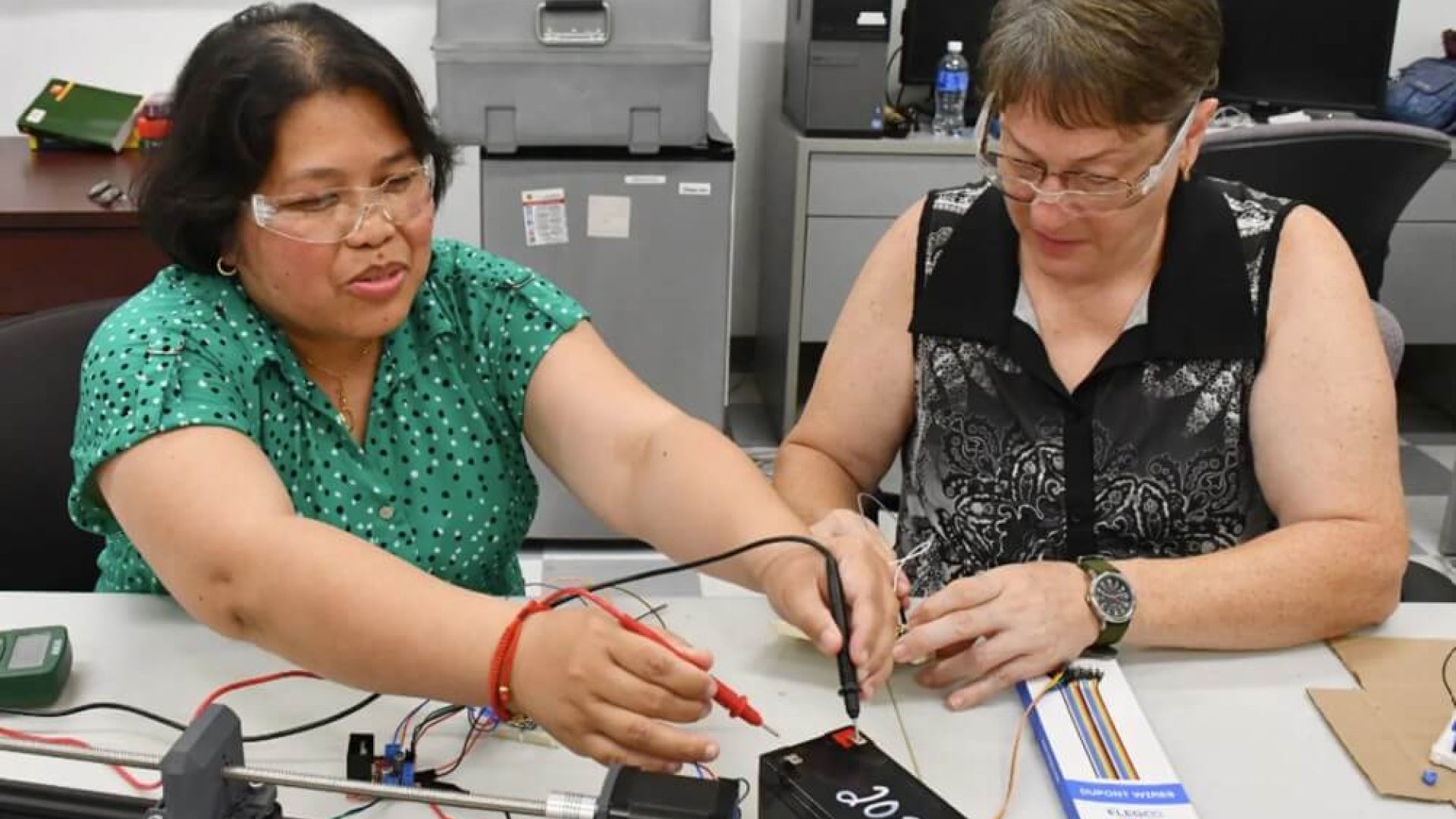 Two women working at a table, one wearing a green shirt and one wearing a black shirt, with school office supplies and equipment on the table and in the background.