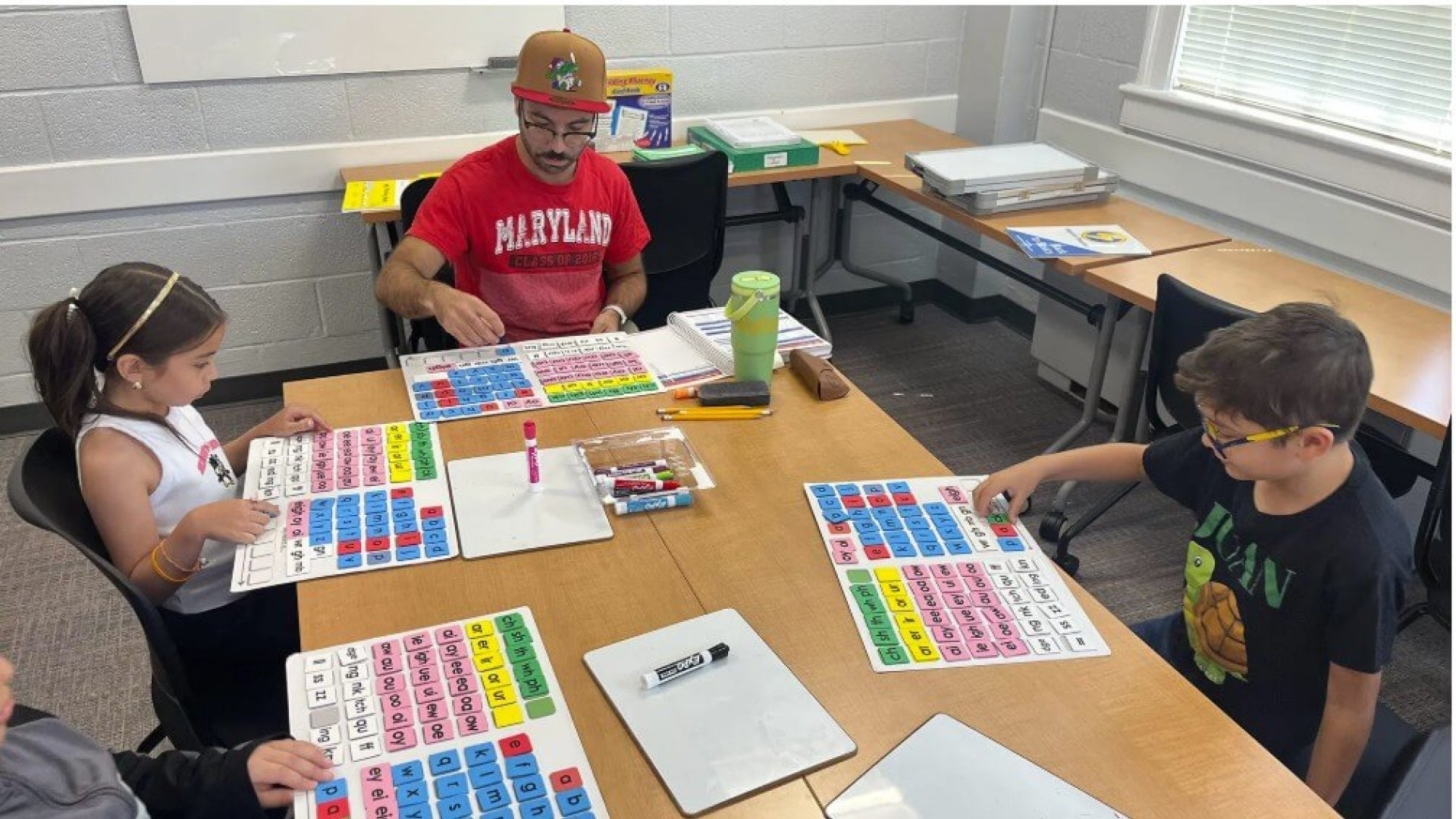 image of man in red shirt that says "Maryland" sitting at a table with kids doing a reading activity. 